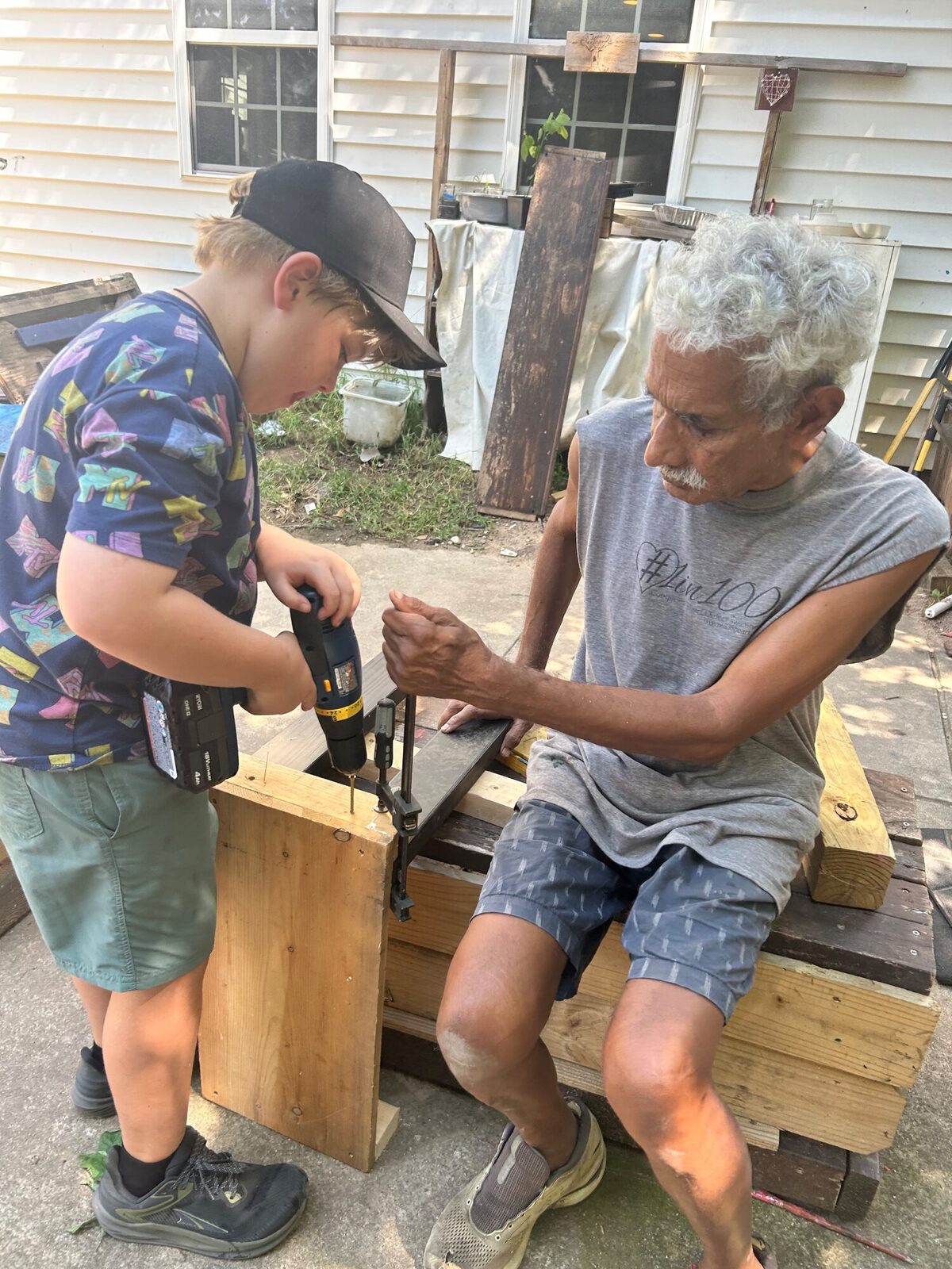 A kid working a hand drill on a plank, with an elder from the community steadying it